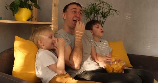 Giovane padre con due piccoli figli si siede sul divano e mangia patatine. Guardando la TV. Illuminazione naturale — Video Stock