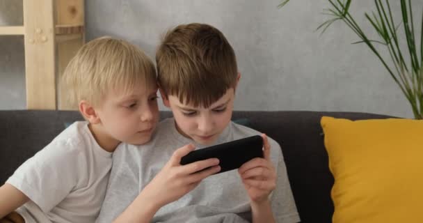 Dos niños jugando en un teléfono inteligente sentado en el sofá. Niños y artilugios. luz del día — Vídeo de stock