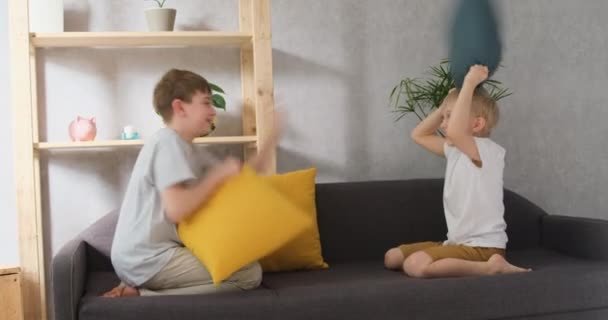 Dwóch wesołych chłopców walczących z poduszkami na kanapie. Światło dzienne widok z przodu slow motion — Wideo stockowe