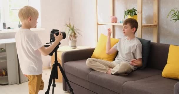 El pequeño camarógrafo rubio le dispara a su hermano en el sofá. Crear contenido de vídeo. Blog de niños. Vista lateral de la luz natural — Vídeos de Stock