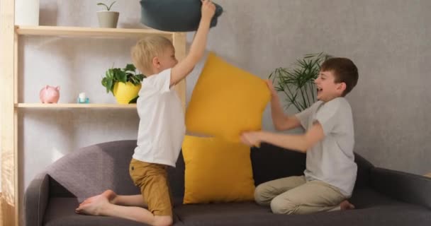 Dois meninos alegres brigando com almofadas no sofá. Visão frontal da luz do dia câmera lenta — Vídeo de Stock