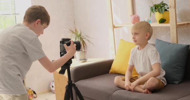 Chlapec zastřelí svého mladšího blonďatého bratra na fotoaparátu doma. Dětský video blog. Přirozený světlý pohled zpomalený pohyb — Stock video