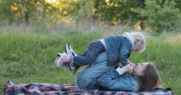 Jonge moeder speelt met haar dochtertje op picknick in het park. Meisje vliegt op mama voeten. — Stockvideo