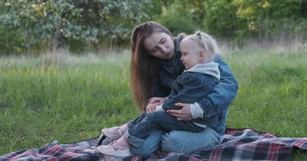 Jovem mãe senta-se com sua filha no tapete de piquenique no parque. Mãe segura menina em seus braços. — Vídeo de Stock