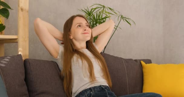 La mujer descansa en el sofá con las manos detrás de la cabeza. Chica piensa en cosas agradables, sueños y sonrisas. — Vídeos de Stock