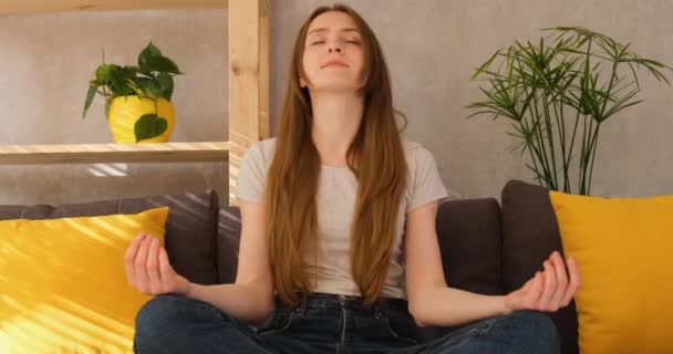 Jovem com cabelos longos está sentado no sofá e meditando. Menina sentada na posição de lótus em casa. — Vídeo de Stock