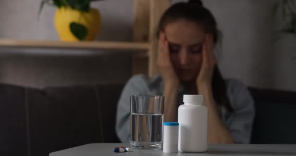 Kranke junge Frau mit Kopfschmerzen mit Tabletten auf dem Tisch im Vordergrund. Mädchen leidet an Migräne. — Stockvideo