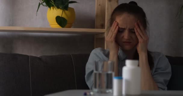Chica sufre de dolor de cabeza y toca las sienes y la cara. Píldoras y agua en la mesa — Vídeos de Stock