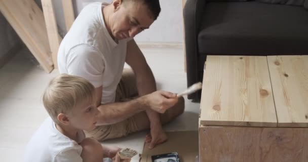 Papá y su hijo pintan mesa en el taller. Padre carpintero enseña a su hijo a trabajar con madera. — Vídeos de Stock