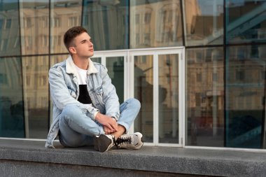 Urban portrait of a stylish guy against the backdrop of the glass facade of the building.H andsome young man resting on the street.