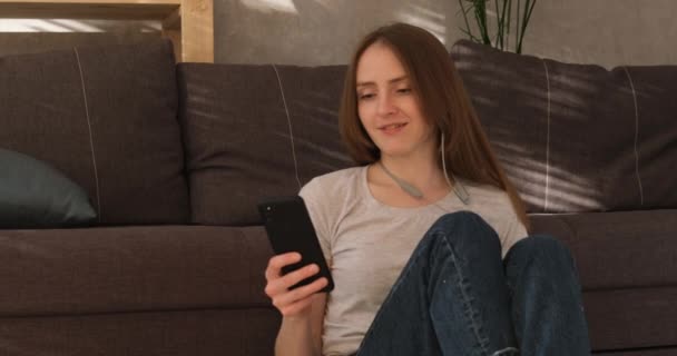 Ung kvinna sitter på golvet i vardagsrummet med telefon i händerna och lyssnar på musik. Flicka sätter på hörlurar och sätter på musik. — Stockvideo