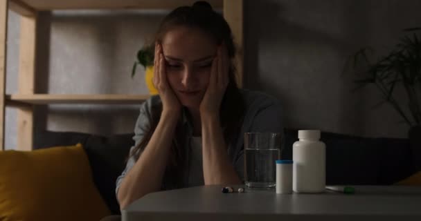 Молода жінка виглядає досить хворим, сидячи біля таблеток. Дівчина з депресією тримає голову в руках . — стокове відео