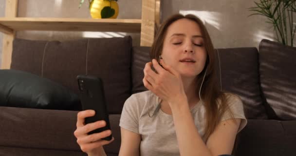 Meisje zet een koptelefoon op en zet muziek aan op de telefoon. Jonge vrouw speelt muziek en knikt met haar hoofd naar de beat. — Stockvideo