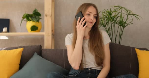 Jovem a falar ao telefone e a sorrir. Menina conversa ao telefone em casa na sala de estar — Vídeo de Stock