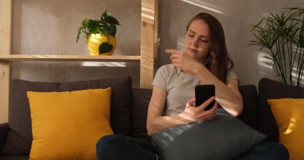 Mujer joven está sentada en el sofá y disfrutando de la música. Chica pone en los auriculares y enciende la música en el teléfono. — Vídeos de Stock