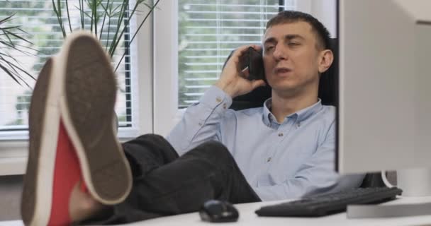 Молодий впевнений чоловік-бізнесмен кидає ноги на стіл. Директор розмовляє по телефону в своєму офісі . — стокове відео
