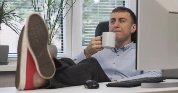 Νεαρός σκηνοθέτης κάθεται στο γραφείο του σε δερμάτινη καρέκλα, πίνοντας καφέ και βάζει τα πόδια στο τραπέζι. Διάλειμμα στη δουλειά, διάλειμμα για καφέ. — Αρχείο Βίντεο
