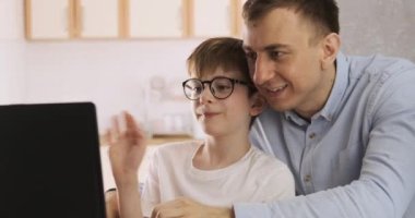 Baba ve oğul evdeki bilgisayar görüntülü konuşma sırasında arkadaşlarına el sallıyor. Ev karantinası, sosyal uzaklık, kişisel izolasyon..