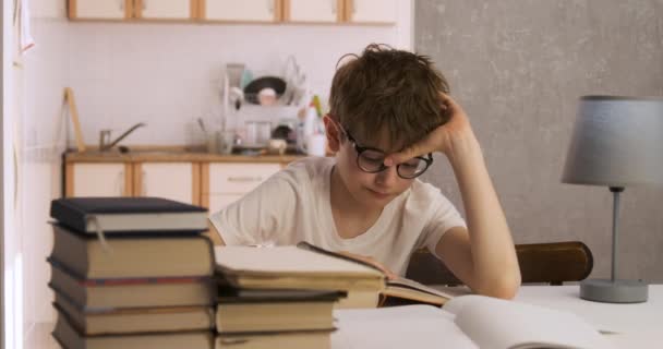 Niño con gafas se sienta en casa entre libros y lee. El colegial hace su tarea. Niño curioso. — Vídeo de stock