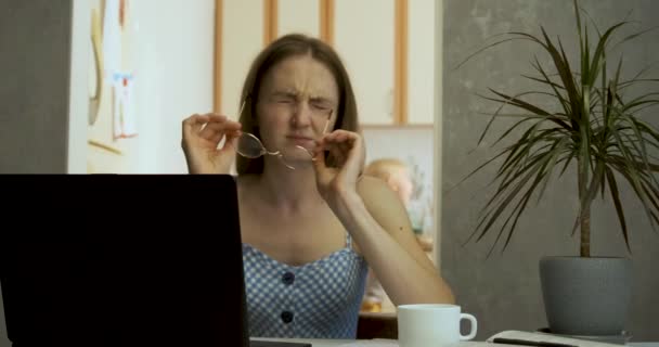 Müde junge Frau nimmt ihre Brille ab und reibt sich die Schläfen, während sie an ihrem Laptop sitzt. Harte geistige Arbeit. Junge Mutter arbeitet zu Hause, während die Kinder hinter ihrem Rücken Lärm machen. — Stockvideo