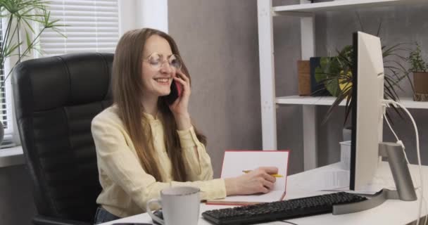 Glada tjejsekreterare vid sitt skrivbord pratar i telefon. Ung affärskvinna på kontoret löser arbetsfrågor. Trevligt telefonsamtal med en affärspartner — Stockvideo