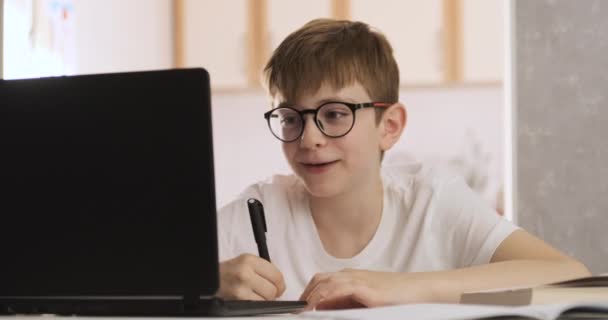 Boy in glasses writes homework while sitting at a table with a laptop. Doing homework. Online training. — Stock Video