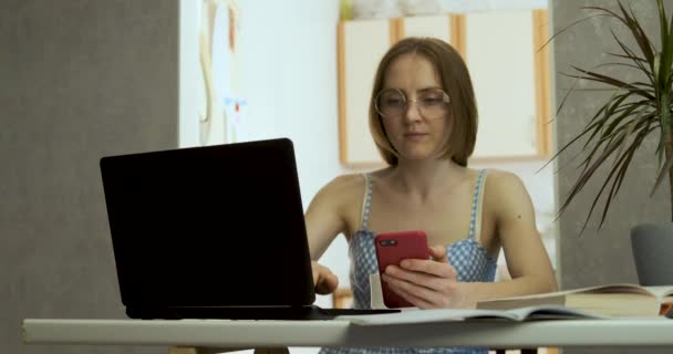 Giovane donna in occhiali con un telefono tra le mani lavora in un computer portatile. Ministero degli Interni, lavoro a distanza. Lavoro in congedo di maternità — Video Stock