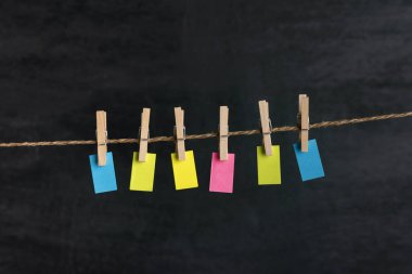 İpte mandallarla asılı çok renkli küçük kağıtlar. Siyah arka plan. Uzayı kopyala Mesajın için yer aç. Not, hatırlatma.