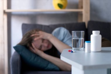 Kadın kanepede yatıyor ve migrenden muzdarip. Baş ağrısından muzdarip kıza karşı ilaç ve bir bardak su.