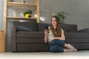 Kız telefon ekranında şaşkınlıkla bakıyor. Evde oturan genç bir kadın, telefonu yerde ve mektupları kontrol ediyor..