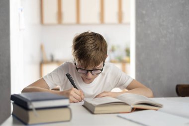 Gözlüklü çocuk ödevini yapıyor. Mesafe öğrenme. Uzaktan eğitim gören çocuklar. Öğrenci ödevlerini bir defterde yazıyor.