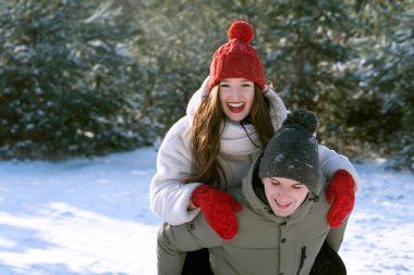 Adam kız arkadaşını sırtında taşıyor. Kış ormanı geçmişi. Genç çift soğuk güneşli bir günde eğleniyor..