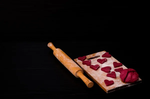 Galletas rojas crudas en forma de corazón y masa sobre tabla de madera con harina, rodillo de madera. Fondo negro. Preparación para el Día de San Valentín —  Fotos de Stock