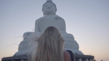 Yavaş çekim: genç sarışın bir kadının Phuket Thailand 'daki büyük beyaz mermer bir Budha heykeline doğru yürüyüşü, rüzgarda savrulan saçlara yakın rüzgarda 120 fps, yaz ve tatil konsepti