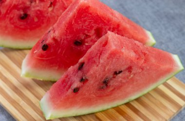 Üç dilim kırmızı karpuz tahta üzerinde duruyor, bambu tahtada, gri bir arka planda.