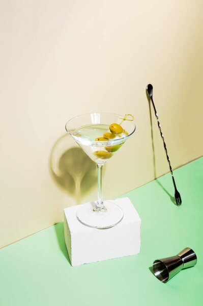 Glass of martini cocktail with green olives on white pedestal. Nearby jigger and bar spoon. Focus on shadows