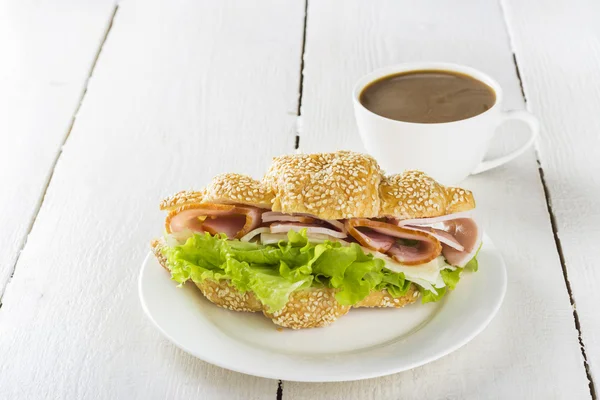 Croissant sendvič, salát, slanina, sýr na bílý talíř a šálek kávy na dřevěné pozadí — Stock fotografie