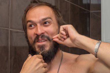 Adam kendini sakalından, iki parmağıyla bıyığından tutuyor ve sakalının yanında makas tutuyor.