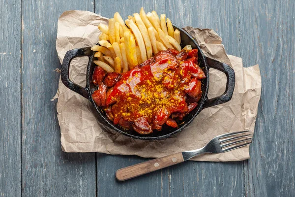 Currywurst avec frites dans une poêle noire sur papier artisanal sur table en bois. Vue du dessus. — Photo