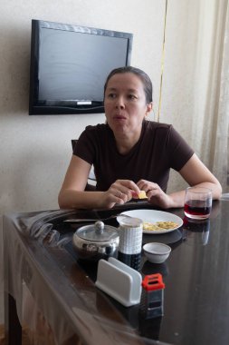 Genç bir kadın mutfakta pencerenin önünde oturmuş kahvaltı yapıyor.