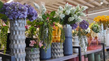 Benzersiz vazolardaki güzel çiçek düzenlemeleri şık bir rafta sergileniyor, parlak bir perakende alanında çeşitli renk ve dokuma çeşitleri yer alıyor.