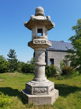 Geleneksel taş fener canlı bir bahçede sergilenir, yemyeşil bitkiler ve açık mavi gökyüzü ile çevrili, sakin dış atmosferi geliştirir.