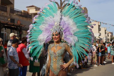 Orihuela Costa, İspanya - 17 Mart 2024: Aziz Patrick Günü geçidi. Renkli bir başlık ve ışıltılı bir kostüm giyen bir sanatçı, festival atmosferinin tadını çıkaran izleyicilerle çevrili bir geçit töreninde duruyor.