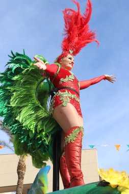 Orihuela Costa, İspanya - 17 Mart 2024: Aziz Patrick Günü geçidi. Renkli kırmızı ve yeşil tüylü dansçı, bir festival sırasında özenle hazırlanmış detaylar ve açık bir gökyüzüne karşı parlak renkler sergileyerek özgüven içinde poz veriyor.