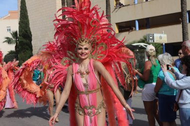 Orihuela Costa, İspanya - 17 Mart 2024: Aziz Patrick Günü geçidi. Tüyleri ve pulları olan renkli bir kostüm giyen kadın, arka planda seyirciler ve palmiye ağaçları ile çevrili bir karnaval geçidi sırasında gülümsüyor.