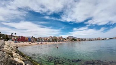 Villajoyosa, İspanya - 03 Eylül 2025: Canlı bir plaj sahnesi, yüzen ve renkli binalar ve atmosferi yükselten açık mavi gökyüzü ile kıyı şeridi boyunca dinlenen insanları yakalıyor.