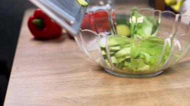 Taze sebzeler doğranır ve ahşap bir tezgahın üzerinde temiz bir cam kaseye dökülür, arka planda kırmızı bir dolma biber ve çeşitli malzemeler düzenlenerek, canlı bir mutfak atmosferi oluşturulur.