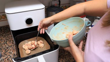 Pembe gömlekli bir kadın, mutfak ortamında bir kase hamur ve mutfak gereçleri ile parşömen kağıdının içine çiğ hamur topları koymak için maşa kullanıyor.