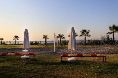 Güneş batarken içi boş voleybol sahası ve Türk otelinde palmiye ağaçları ve deniz var..