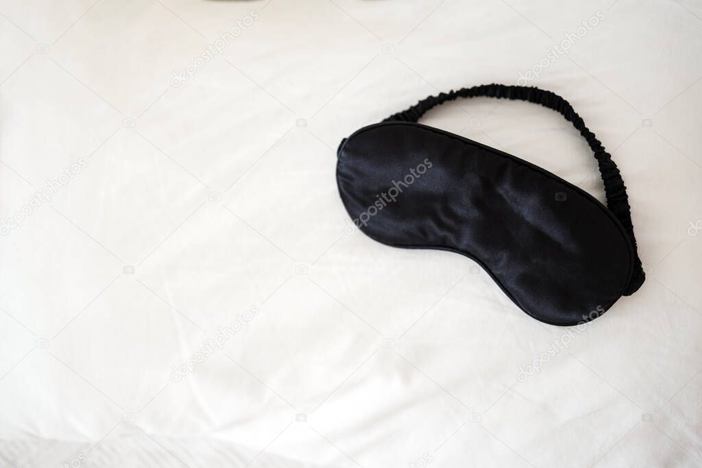 Black satin-silk sleep mask on white bed linen background, no people. Copy space. Comfortable relaxing sleep concept.